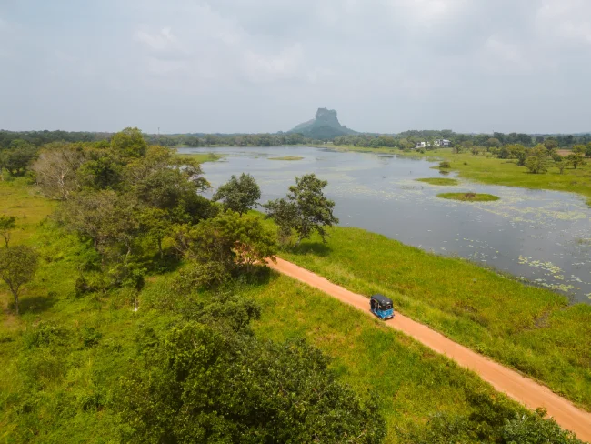 A blue tuktuk driving along a dirt road next to a lake with Lion's Rock in the distance in Sigiriya, Sri Lanka