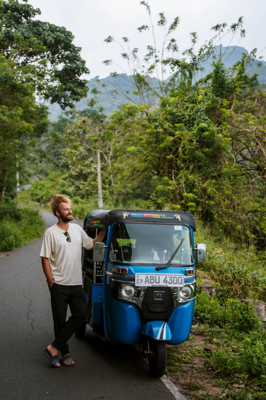 A man stands next to a blue tuktuk on a narrow paved row lined with thick jungle on both sides