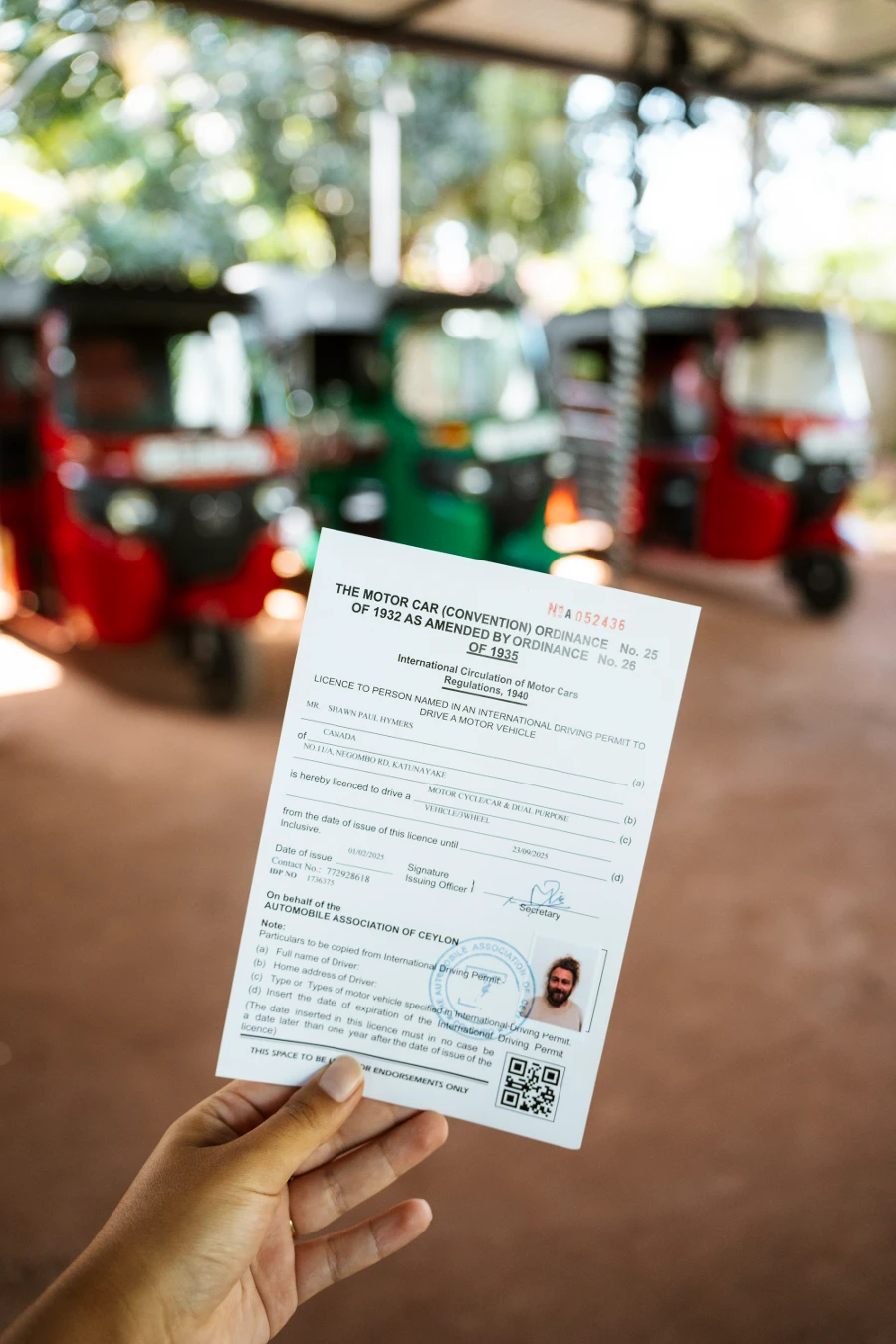 A hand holds up a tuktuk driving license in front of a line of red and green tuktuks