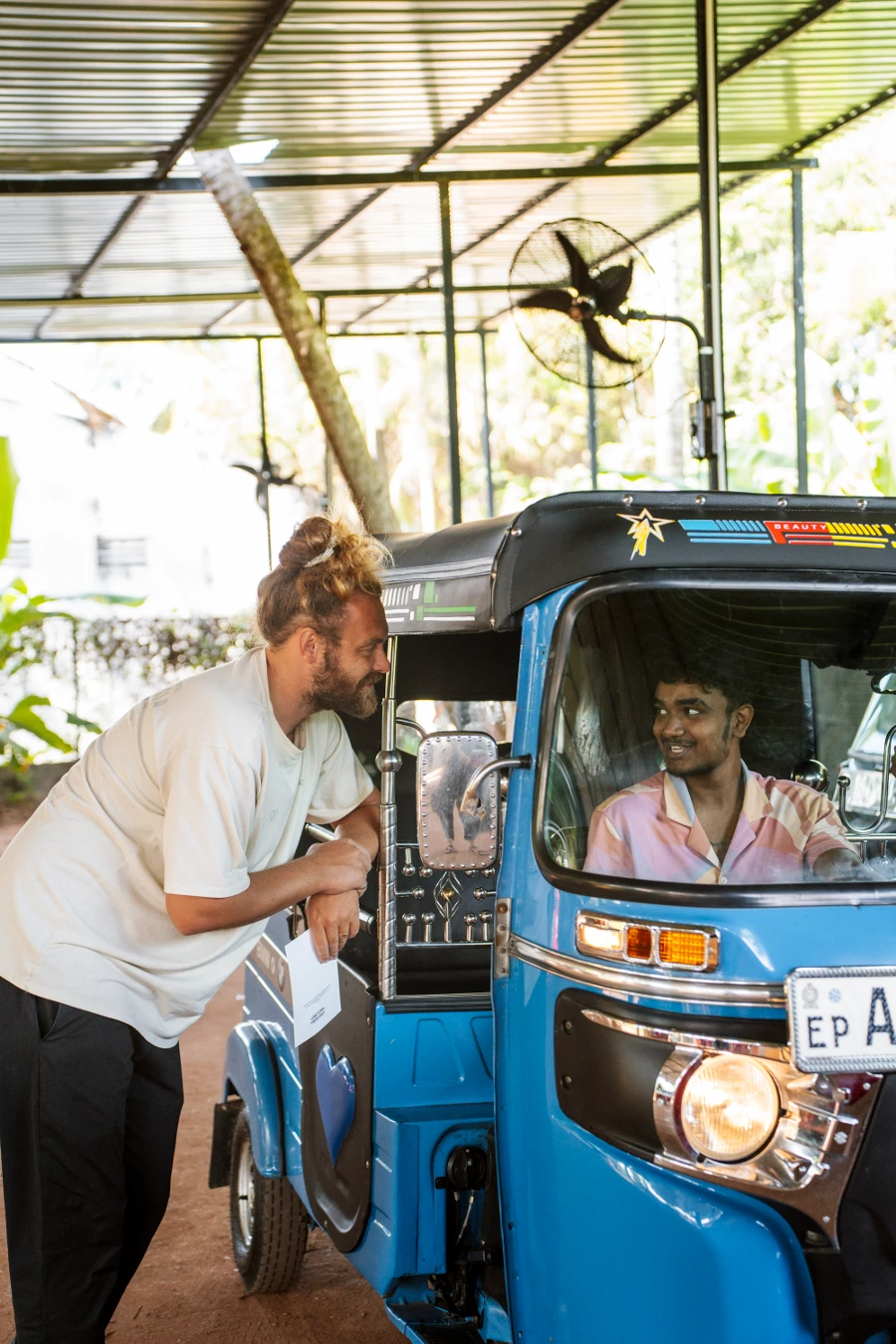 A man holding papers leans in to talk to another man sitting inside a blue tuktuk