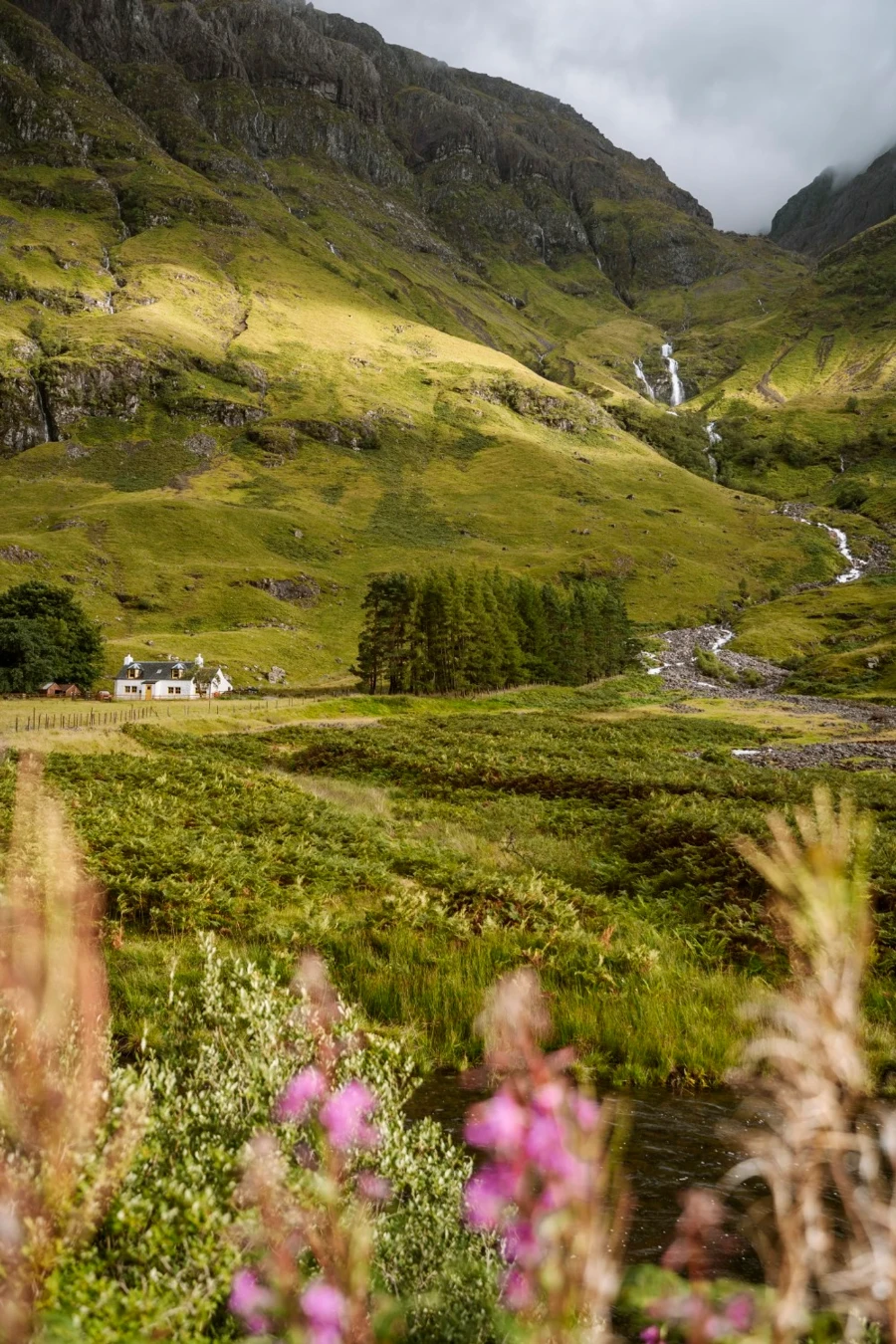 Two waterfalls drop along side a green mountain and pass by a white cottage in the valley below in Glencoe, Scotland