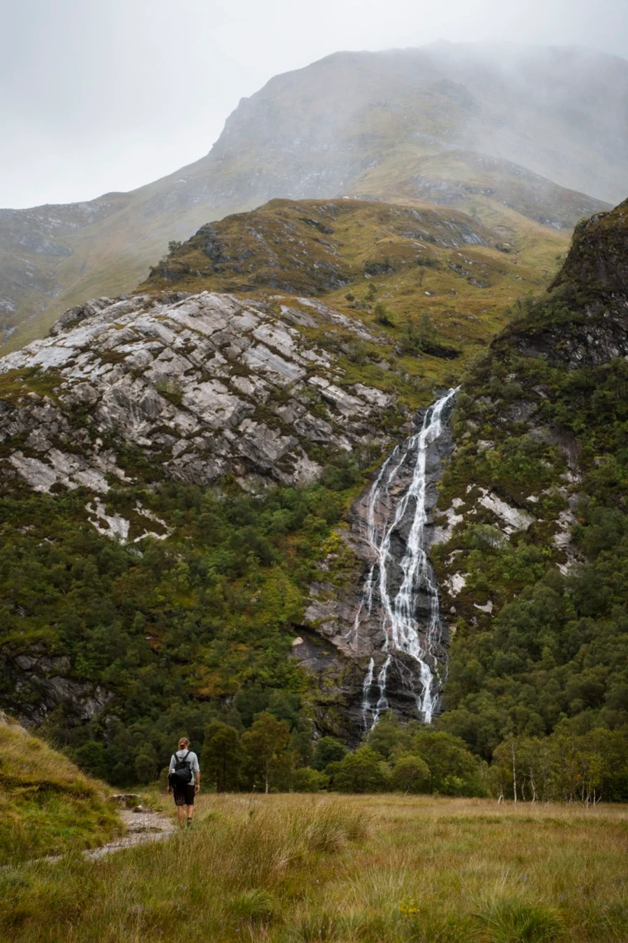 A man walks along a narrow path towards the multiple streams of water pouring over a cliff face known as Steall Waterfall