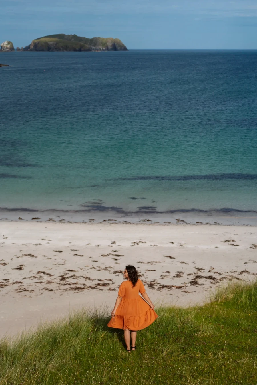 A woman in an orange dress stands on the edge of a grassy cliff looking out to a white sand beach