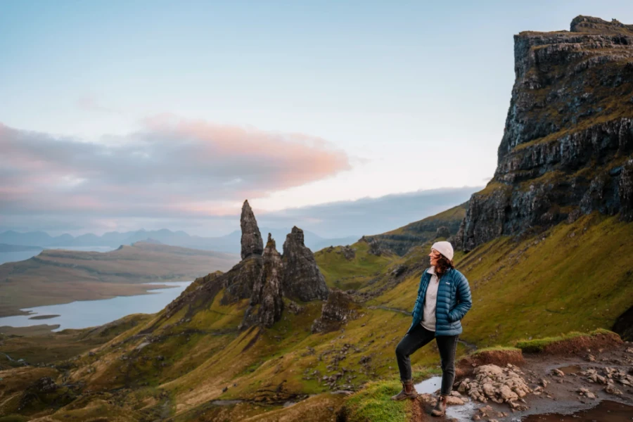 A woman stands at the edge of a cliff with the rocky outcrop known as The Old Man of Storr in the distance