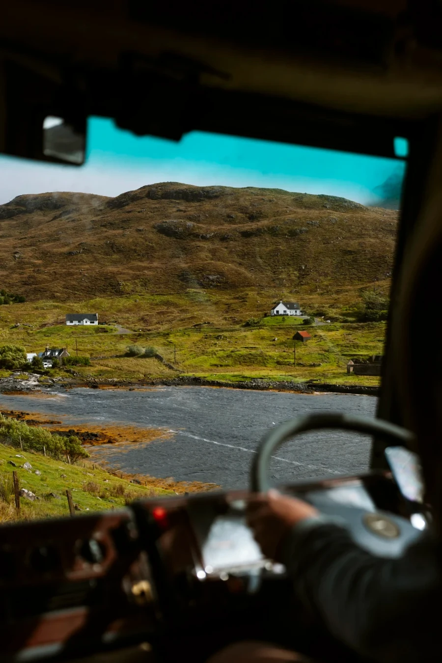 A man grips the steering wheel of a bus through the windshield of which you can see a narrow single track road leading to a small cluster of white cottages