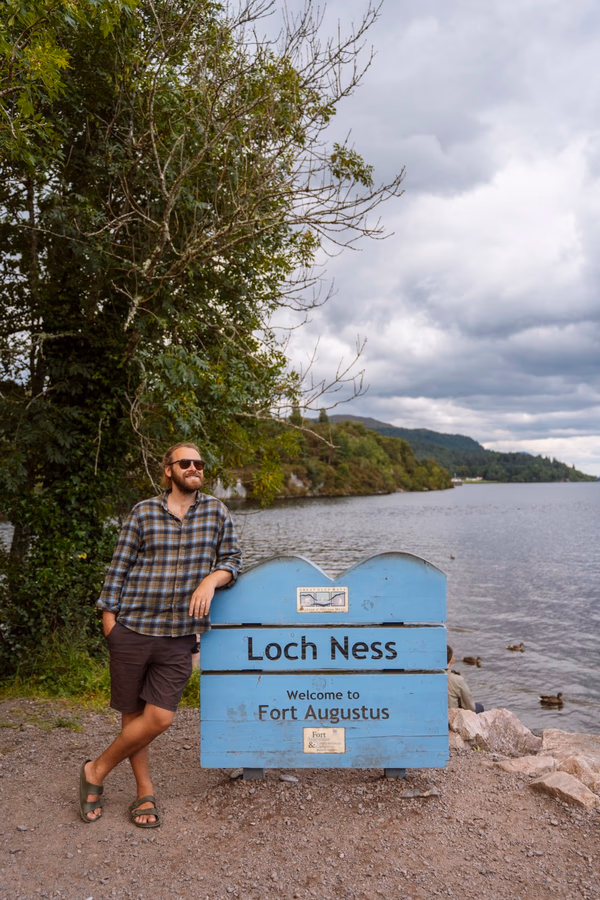 A man leaning against a sign next to a body of water that reads Loch ness