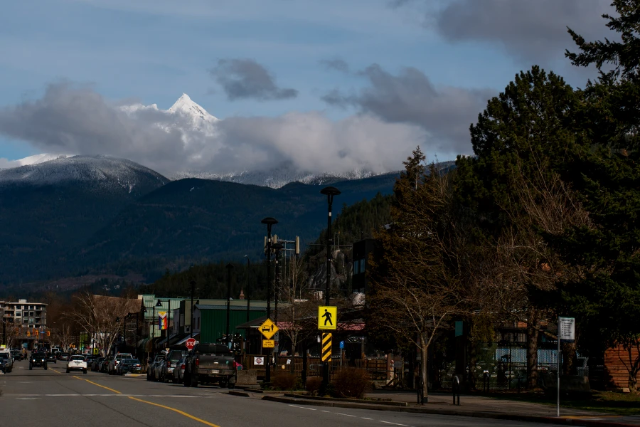 Cars drive down a tree lined steets in downtown Squamish with snowy peaks filling the sky in the distance