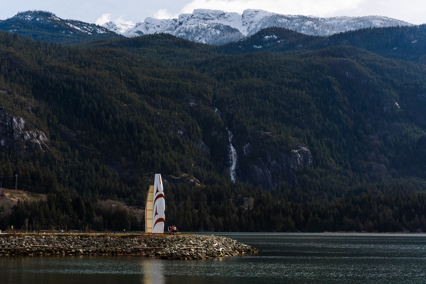 A monument sits on a rocky outcropping that sticks into the Howe sound with Shannon falls falling down a forested mountainside in the distance