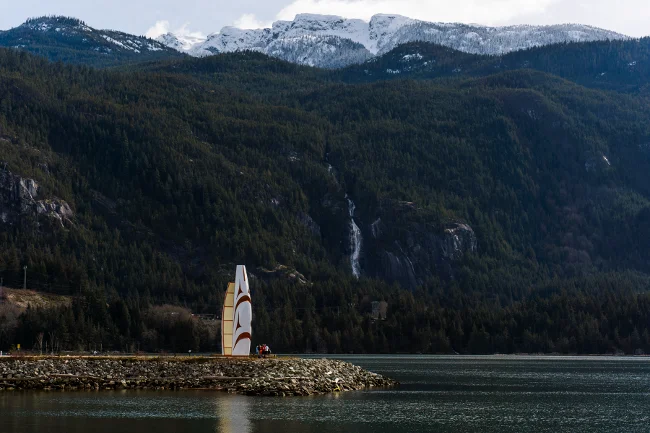 A monument sits on a rocky outcropping that sticks into the Howe sound with Shannon falls falling down a forested mountainside in the distance