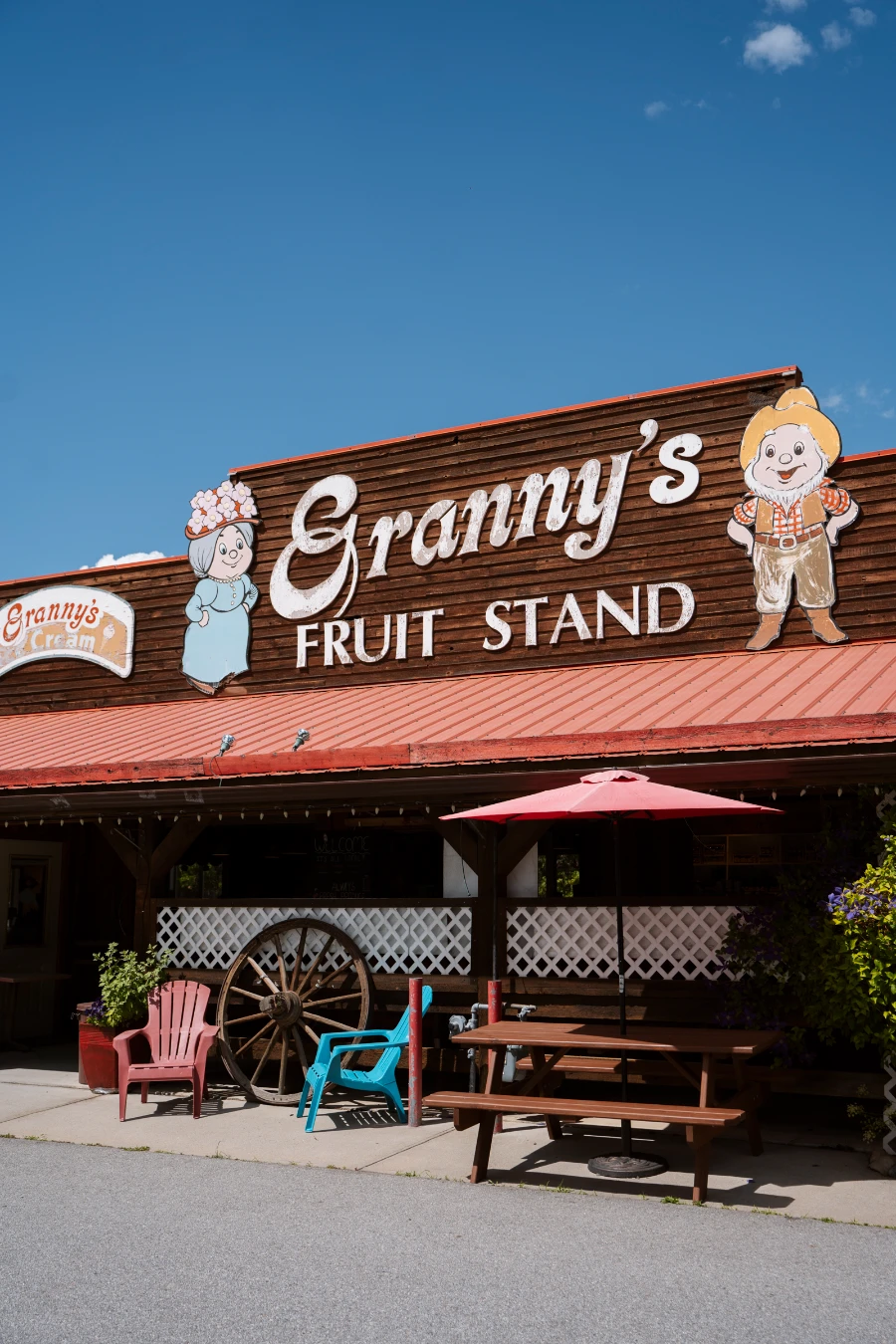 A picnic table and 2 Muskoka chairs sit on a sidewalk in front of a single-story large building that has a sign with 2 cartoon characters standing on either side of large text that reads ‘Granny’s Fruit Stand’