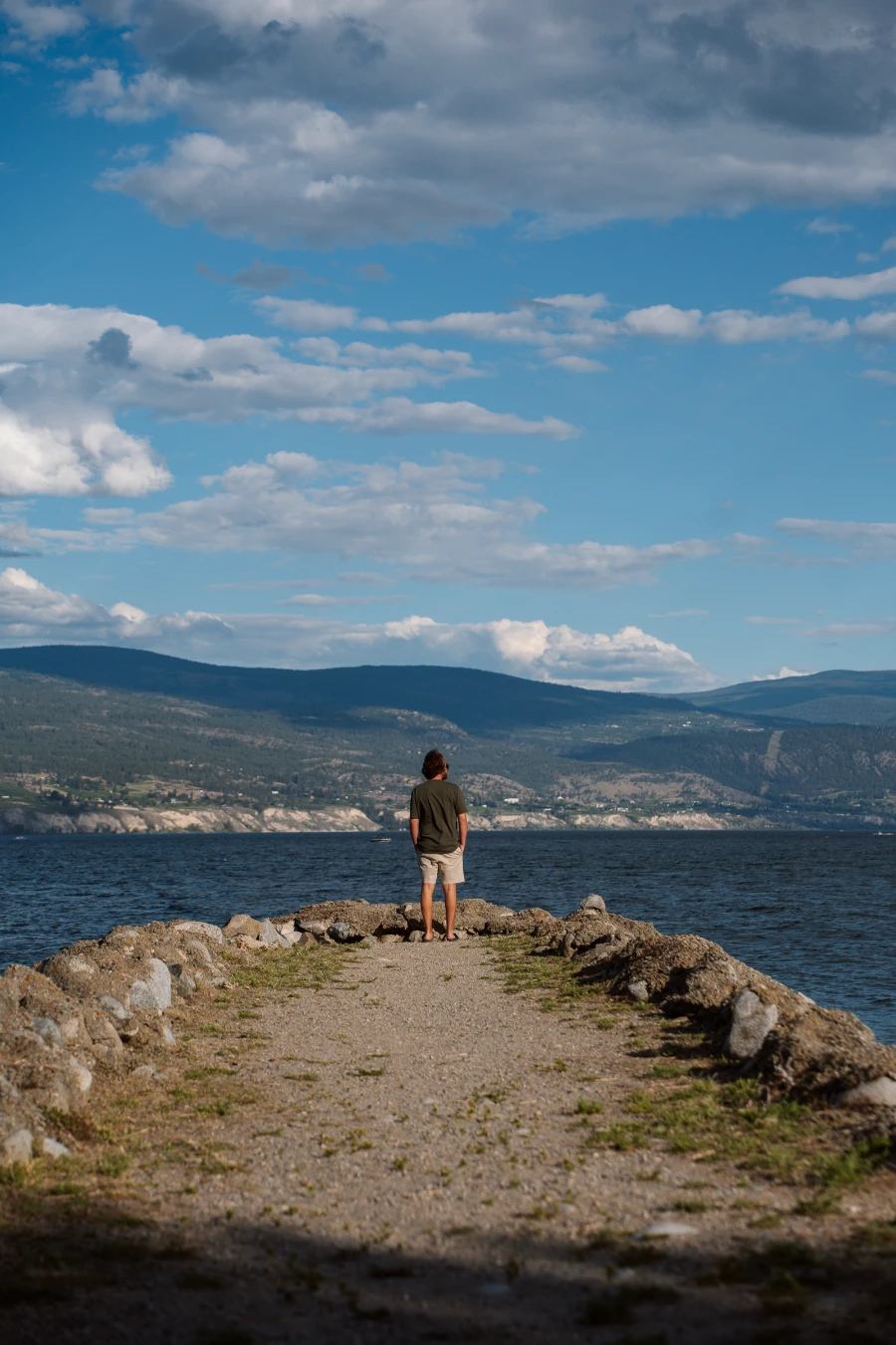 A man stands at the end of a dirt path made into a natural stone pier that juts out into the waters of Okanagan Lake at Gordon Beggs Rotary Beach