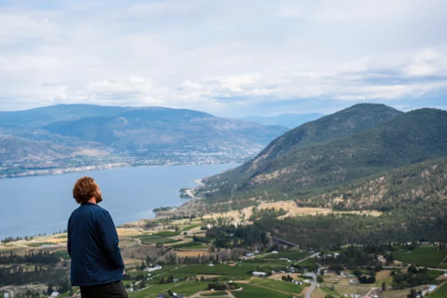 A man stands at the top of Giant’s Head Mountain with the sweeping landscape of farmlands, forested mountains, and Okanagan Lake beneath him