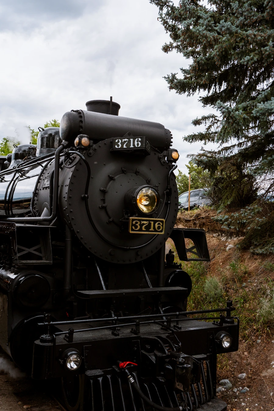 The front of a black-painted old-style steam-powered train engine sitting on the track next to a dirt ditch that is lined by trees