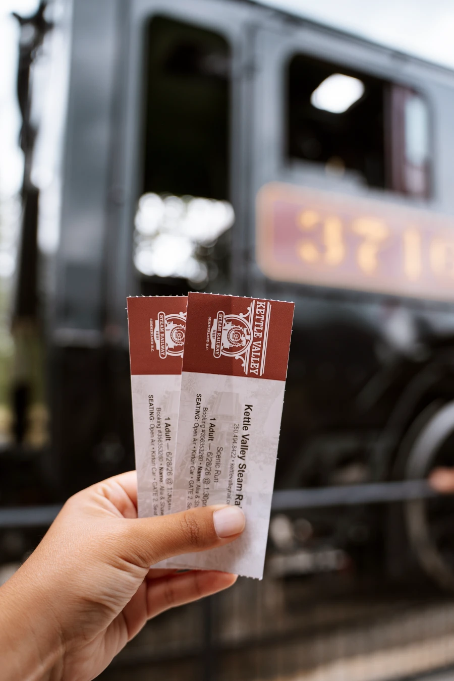 A hand holds two paper tickets up in front of a large black steam train engine car.