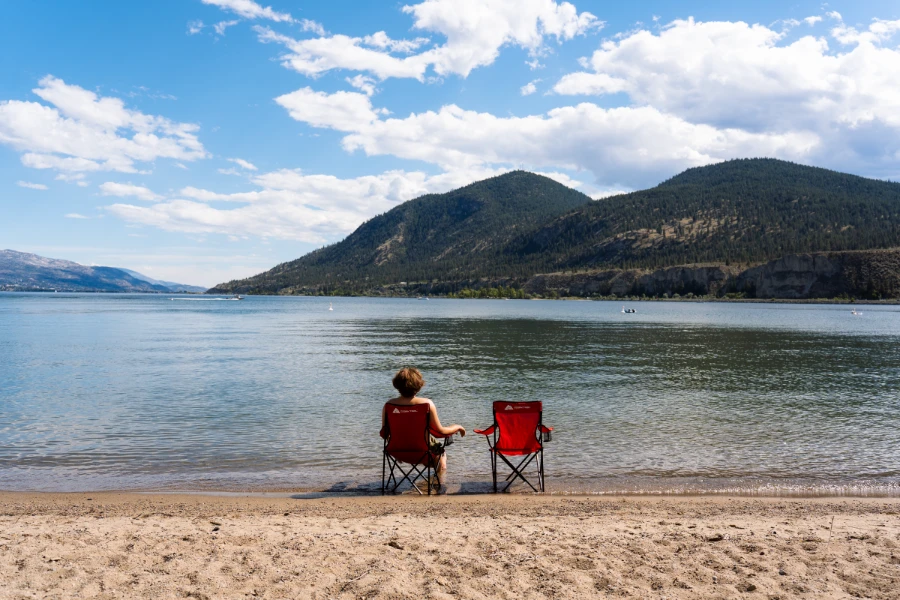 Two red camp chairs sit at the edge of a calm lake at Sun Oka Beach in Summerland, half in the sand and half in the water, with a man sitting in one and the other empty