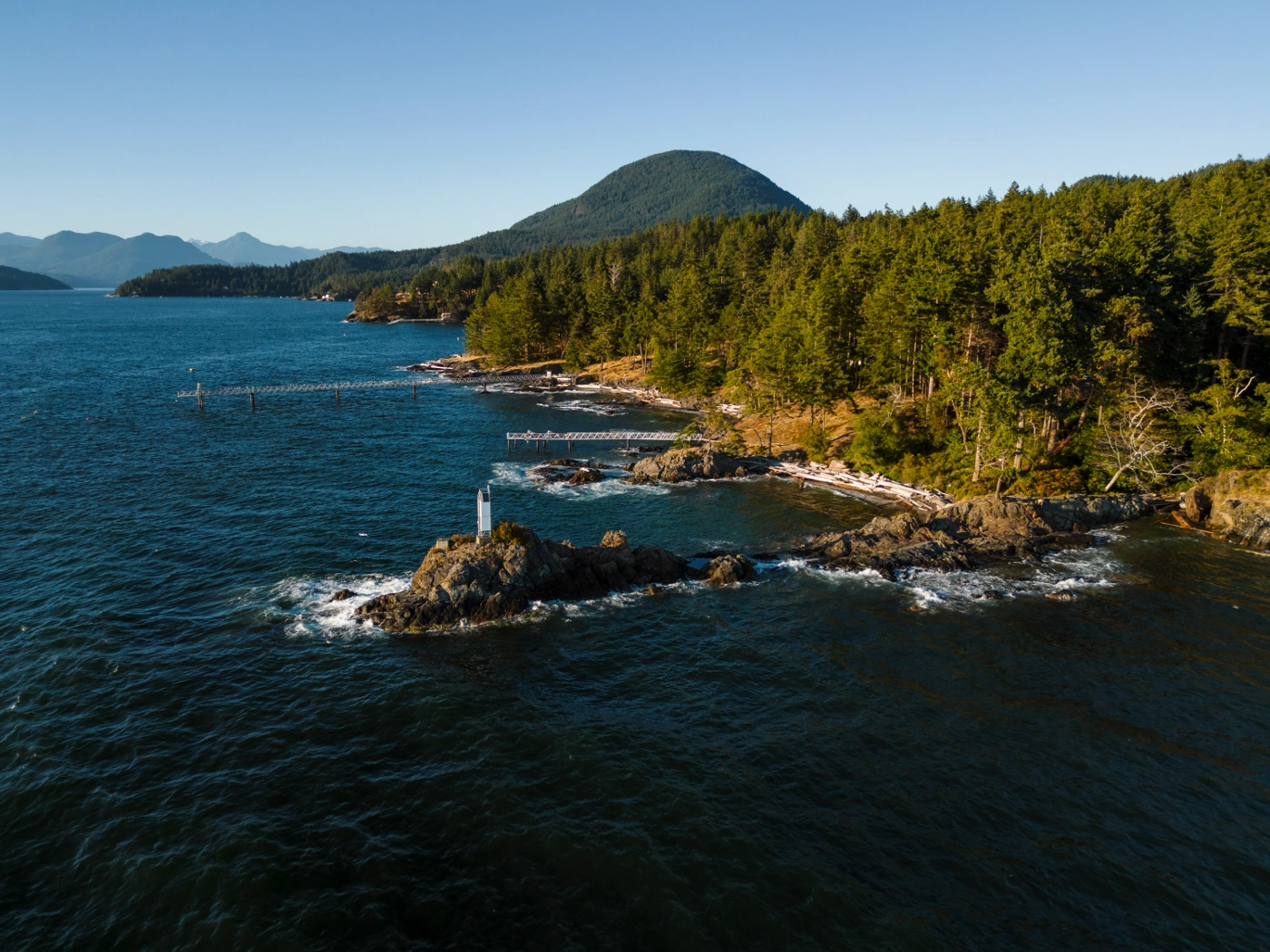 a ttree-lined shore sticks out along a rocky out crop with a small white lighthouse perched at the end on Bowen Island, BC