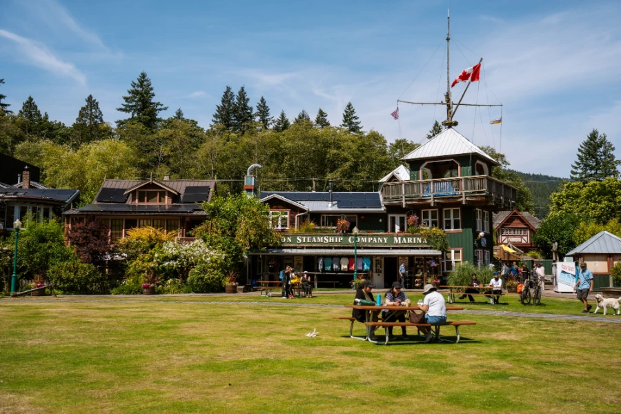 a few small groups sit at picnic tables on the grass outside of an old green building that reads Steamship Company Marina on Bowen Island, BC