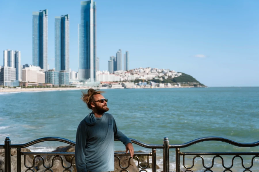Man standing leaning on a metal railing in Blueline Park with large glass towers lining a beach across the water behind him
