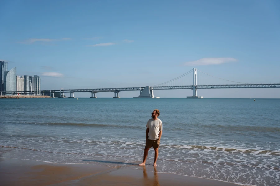 Man stands ankle deep in calm ocean water at Gwangalli Beach with Gwangan bridge crossing the water in the distance behind him