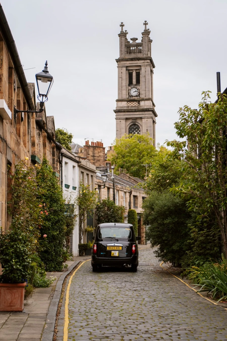 A black taxi cab drives down a narrow cobblestone street in Stockbridge