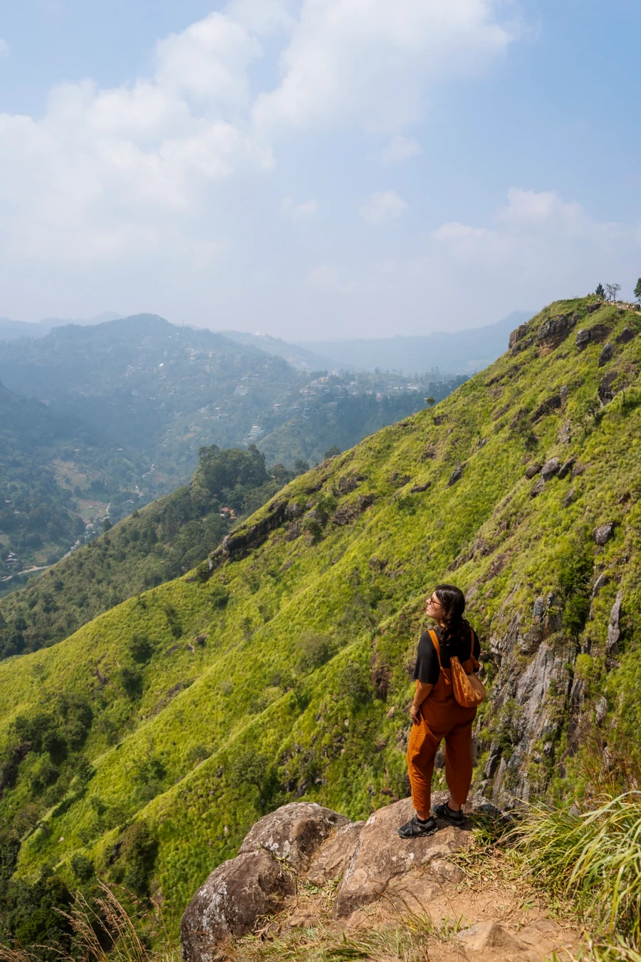 A woman stands on a rock looking down at a green valley below from Little Adam's Peak