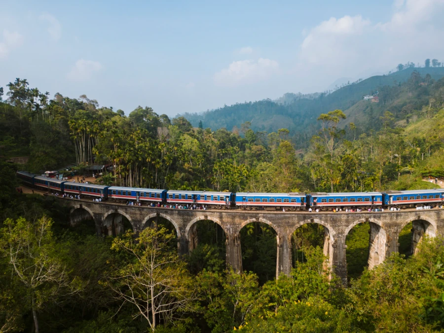 A long blue and red train sits parked on a stone nine arch bridge that spans over a dense jungle below
