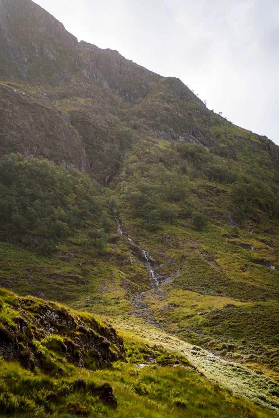 A waterfall flows next to a steep trail that leads up into the mountains to Lost Valley (Coire Gabhail) near Glencoe, Scotland.