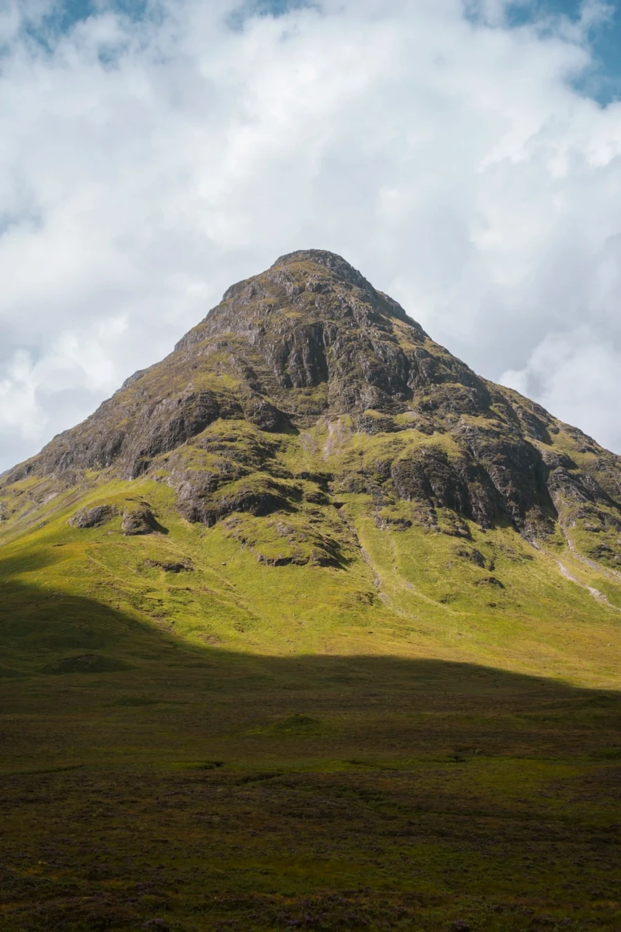 A large shadow is cast over the base of the mountain named Buachaille Etive Mòr while its rocky peak is lit by bright sun
