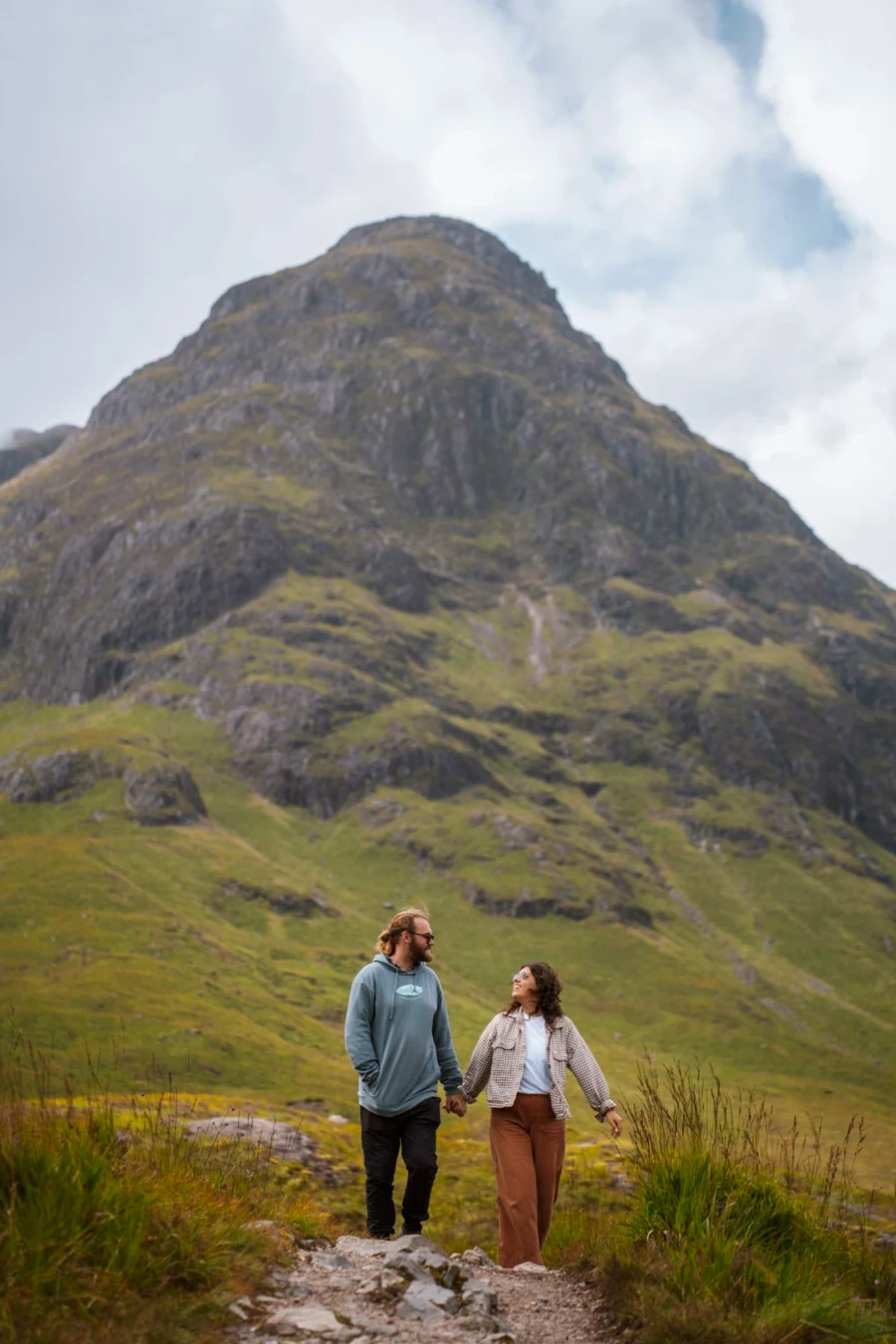A man and a woman walk hand in hand in front of the mountain named Buachaille Etive Mòr