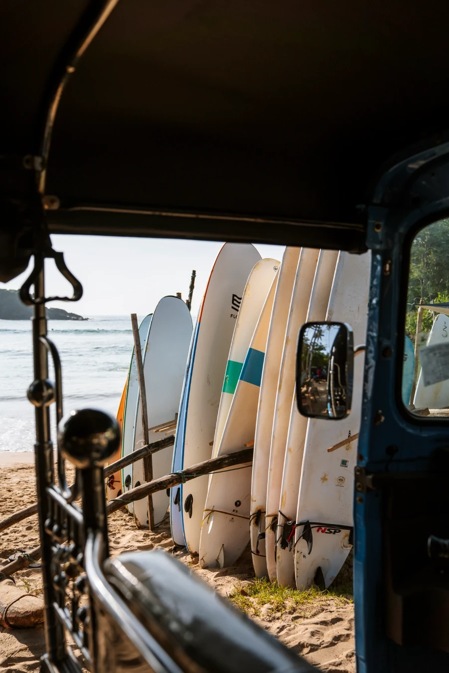 A line of surfboards standing vertically on a sandy beach framed by a tuktuk