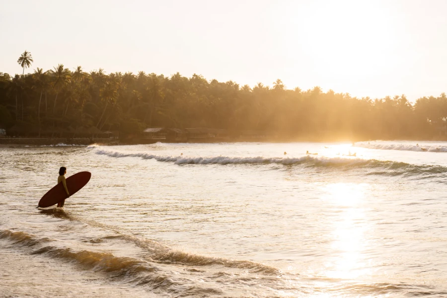 Man holding a surf board standing knee deep in water looks out to the waves hitting Hiriketiya beach at sunrise