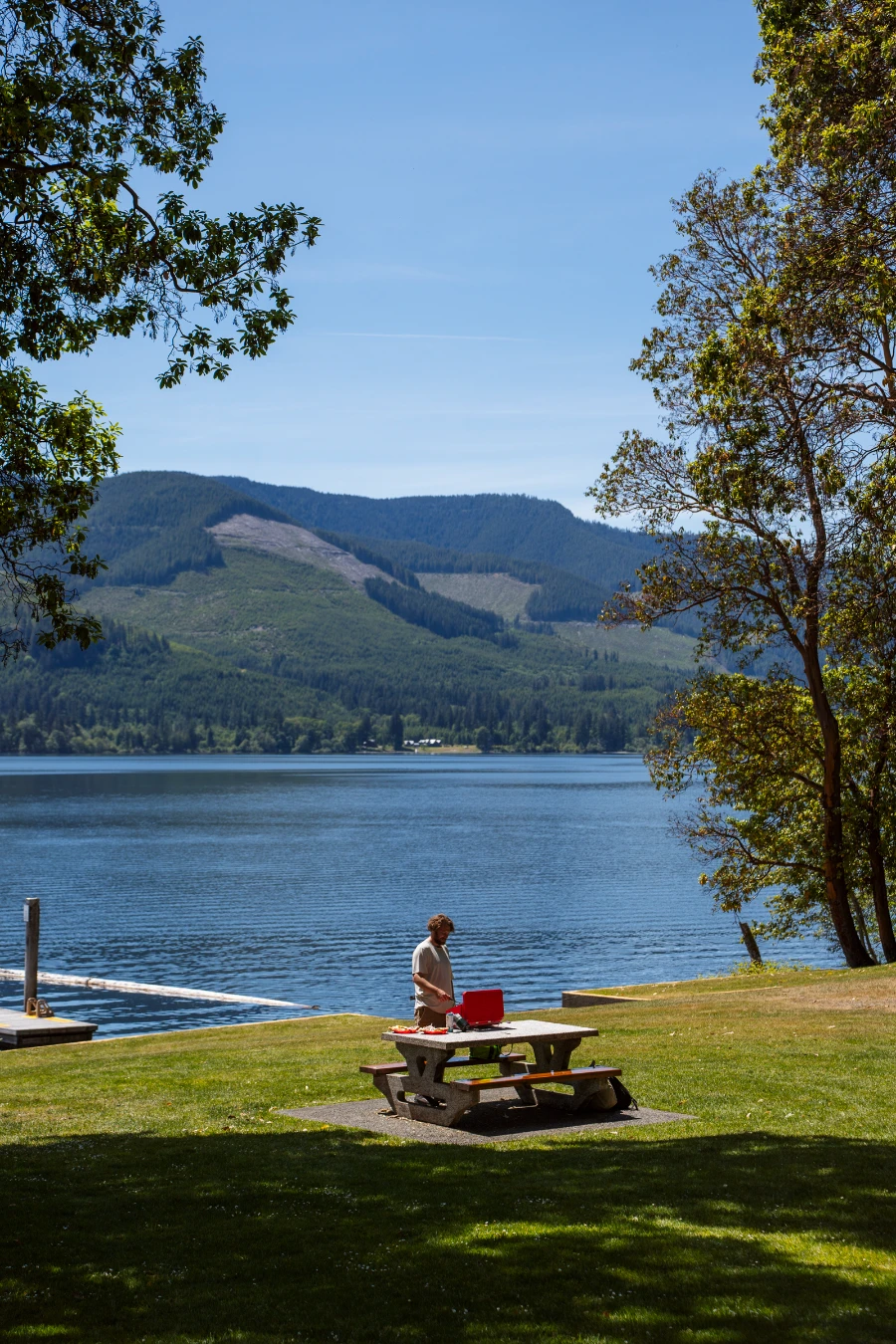 A man cooking on a small red camp stove set up on a picnic table in a grassy park on the edge of Cowichan Lake