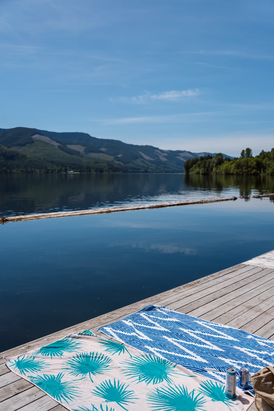 Two beach towels laid out on a wooden dock on Cowichan Lake, with several floating logs in the water marking out a bordered swimming area