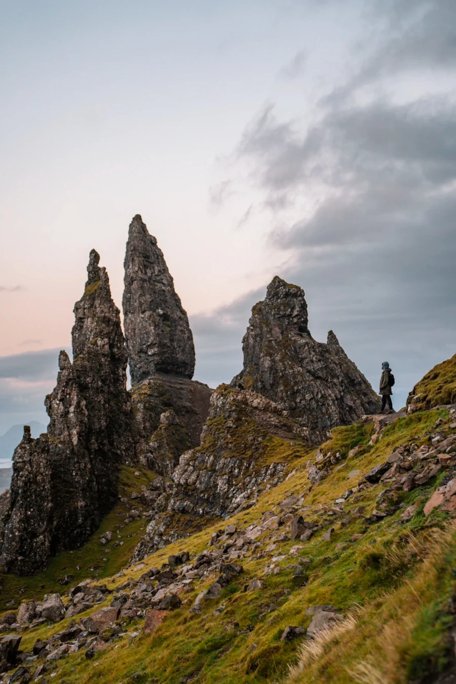 A man standing in the distance looking small next to the massive stone pillars of the Old Man of Storr