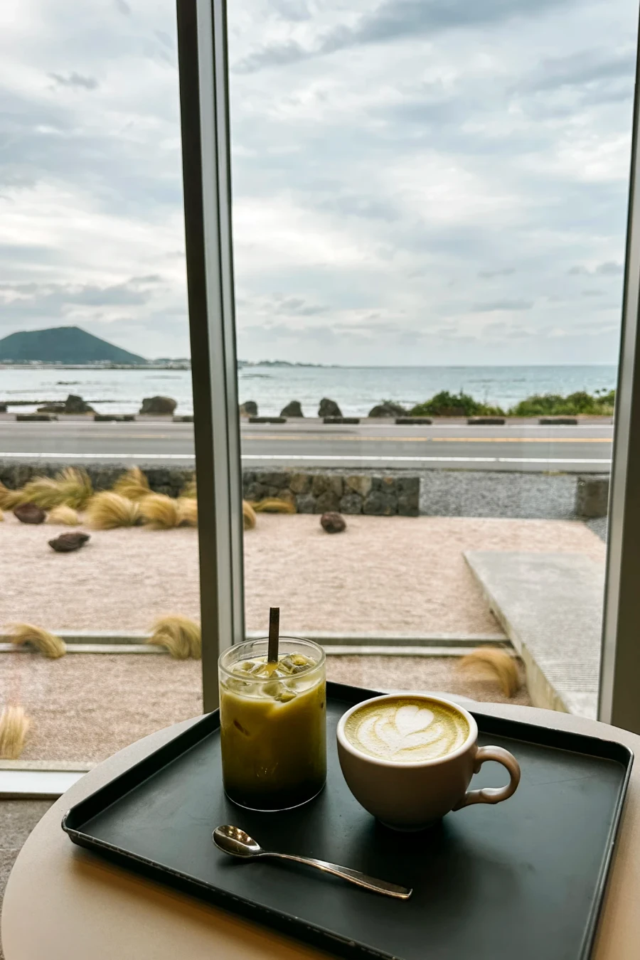 Two well crafted matcha drinks placed on a black tray in front of a window at Cafe Orrrn that looks out to the coastline of Jeju Island