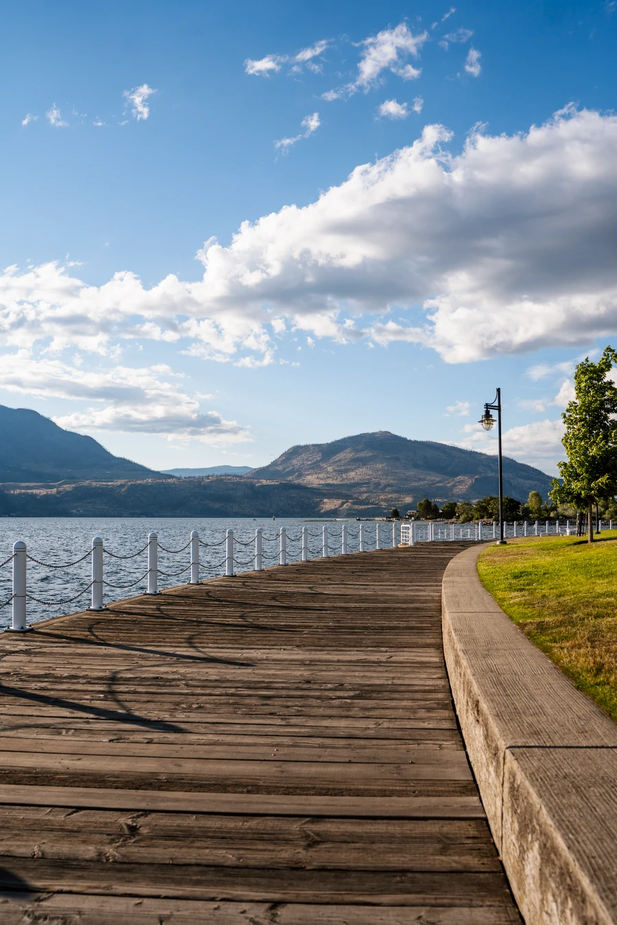 A curved wooden boardwalk winds past the calm waters of Okanagan Lake and a manicured lawn