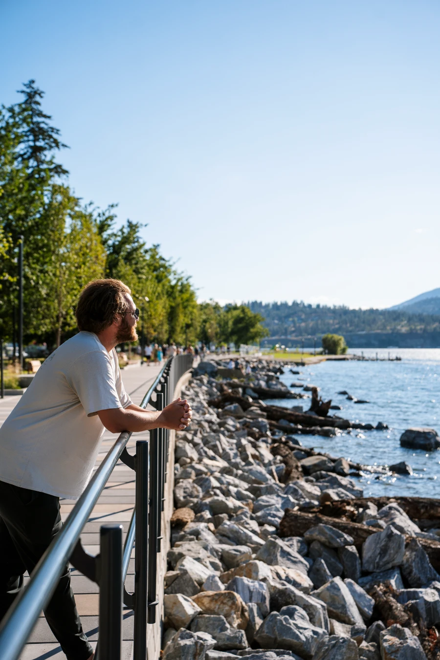 A man leans on a metal railing that runs along the Kelowna Waterfront next to a large pile of rocks that line the water’s edge