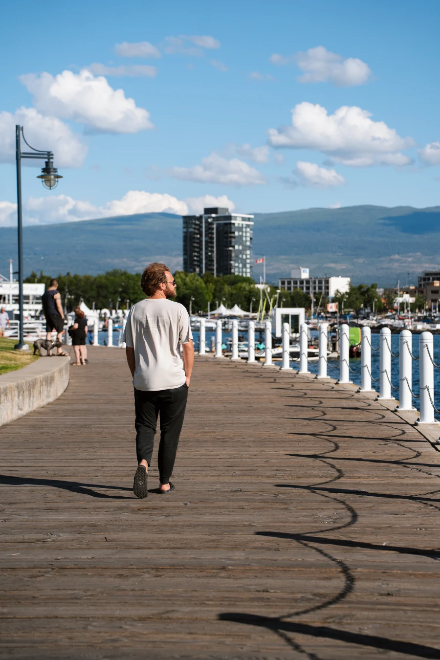 A man walks along a flat and curved wooden boardwalk along Kelowna’s Waterfront with the lake and several high-rise buildings in the distance
