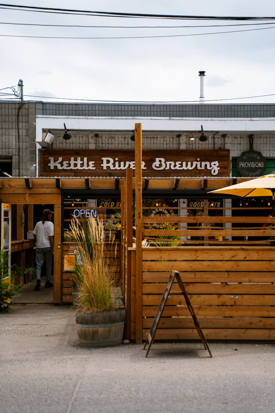A street-side patio closed off by stained wooden fences sit infront of a small building with a wooden sign that reads ‘Kettle River Brewing’