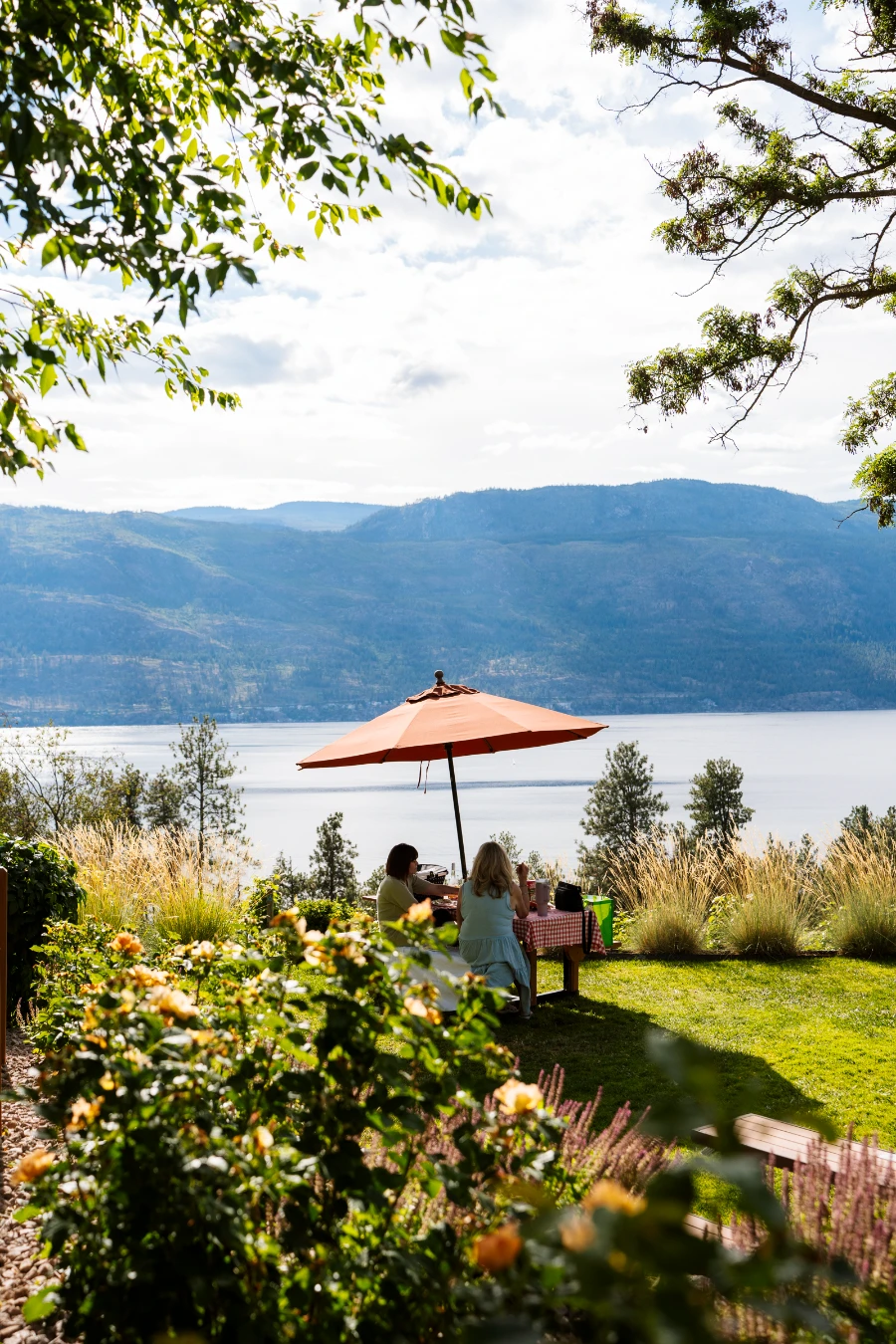 Lush bushes frame a manicured lawn where two women sit at a picnic table in the shade of a large umbrella with Okanagan Lake in the distance