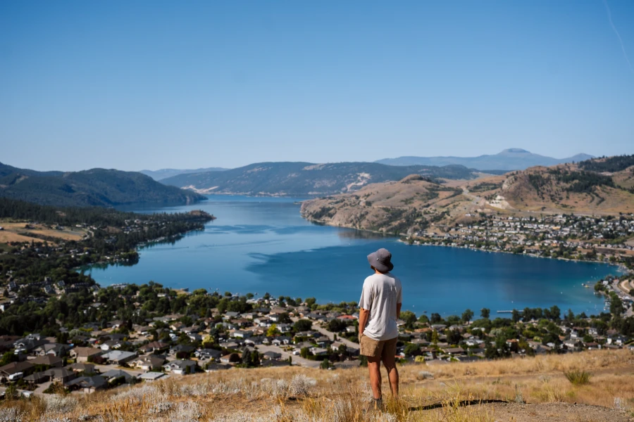 A man stands on a hill of short and dry yellow grass looking out at Kalamaka Lake and a collection of suburban homes below