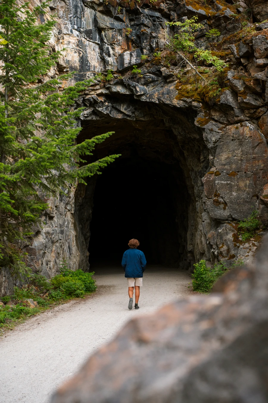A man walks along a wide gravel trail through the Myra Canyon, heading into a large pitch-black tunnel
