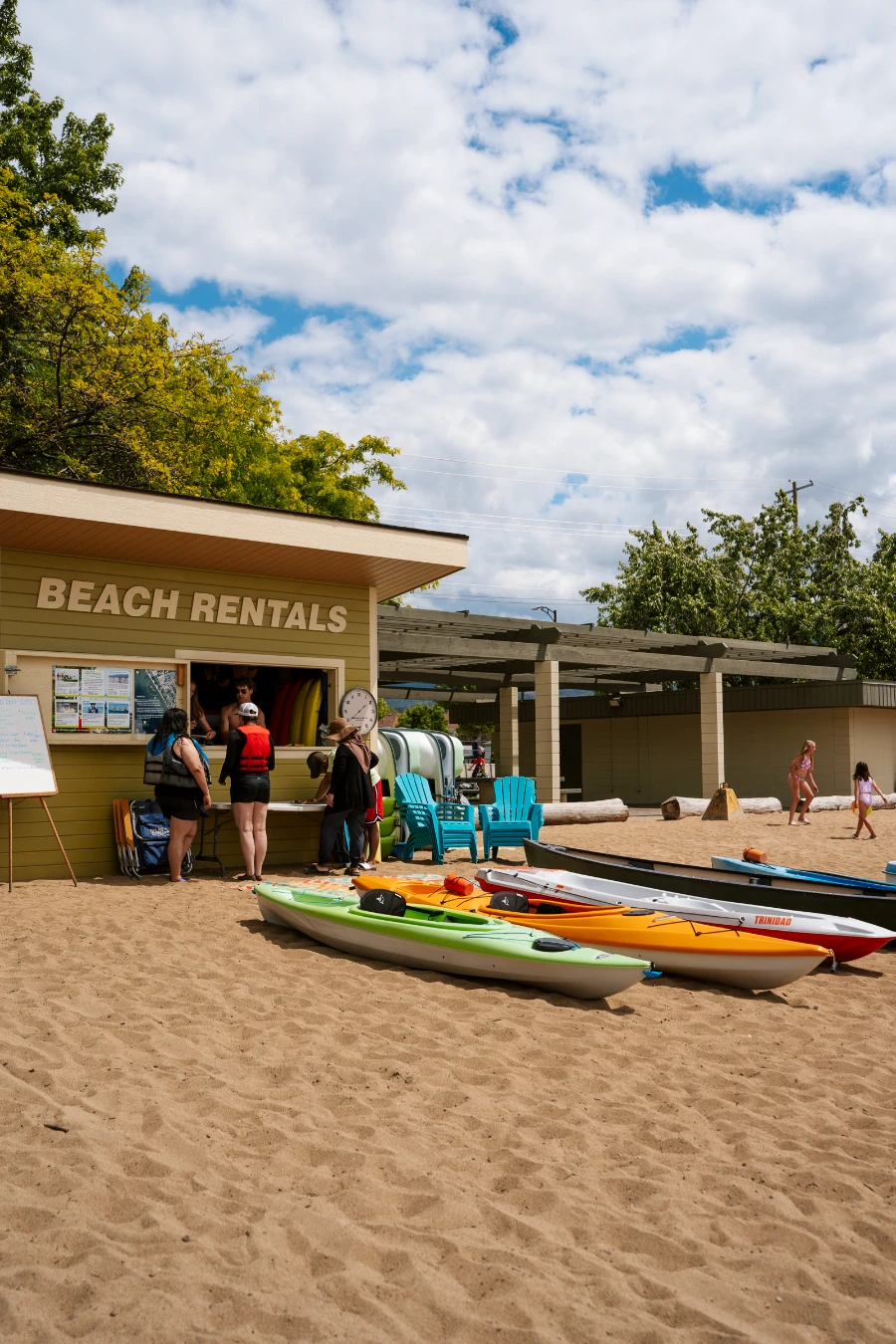 Several kayaks lay in the sand at a Kelowna city beach in front of a building with a line of people and a sign that reads ‘Beach Rentals’