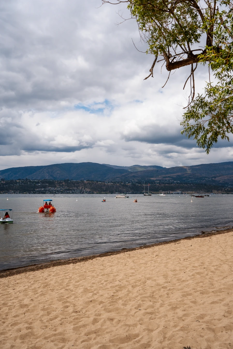 Sandy shores of a city beach in Kelowna with several unique watercraft navigating the calm waters of Okanagan Lake in the distance.