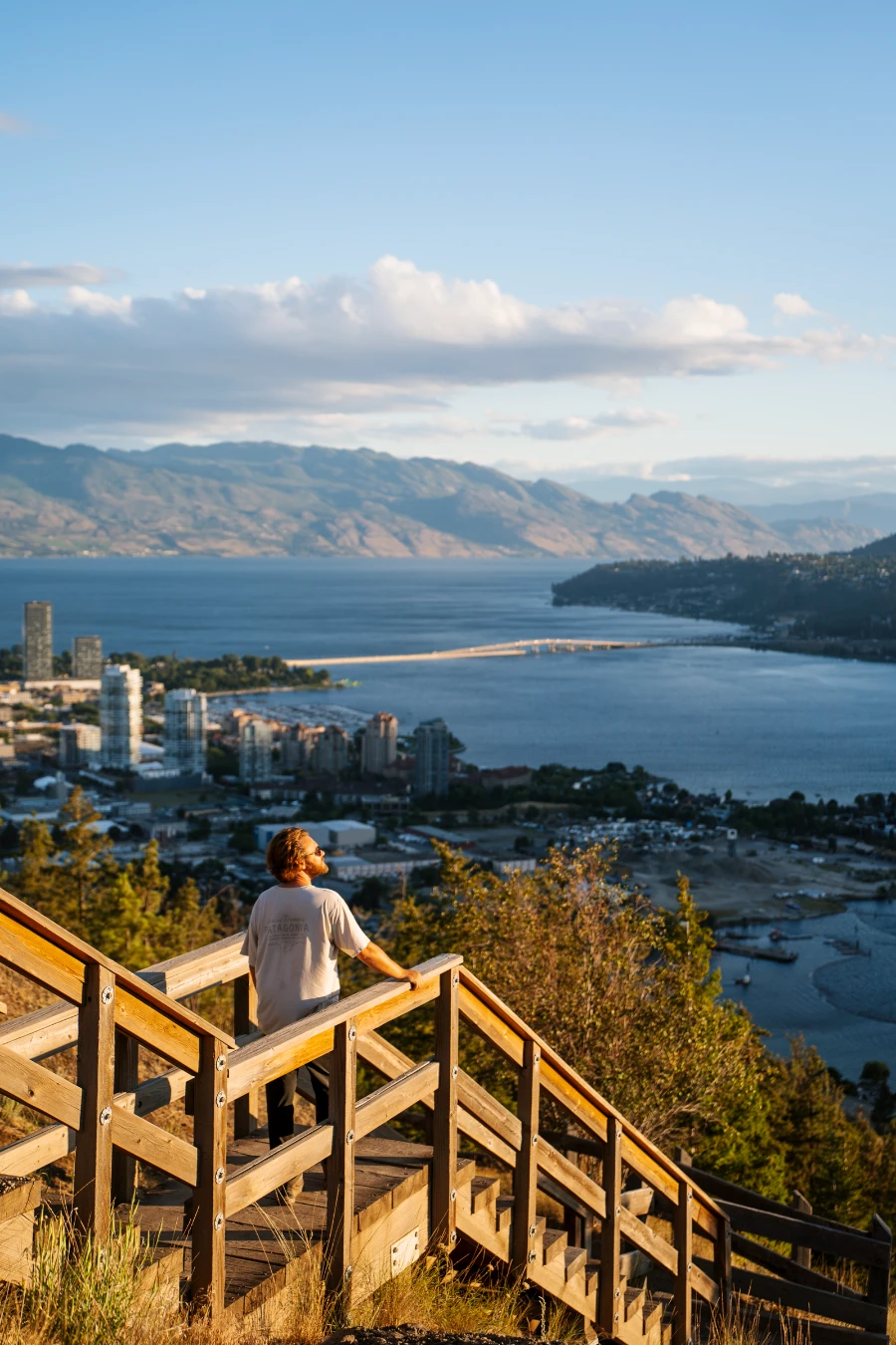 A man stands on a wooden staircase at the top of Knox Mountain looking down at the city of Kelowna and Okanagan Lake below