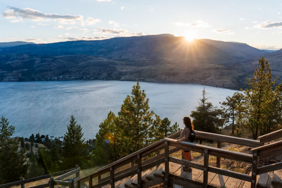 Sunflare shoots in all directions as the sun tucks behind a distant mountain across Okanagan Lake and streams between the sloped ceiling and large railing of a gazebo built on top of Knox Mountain