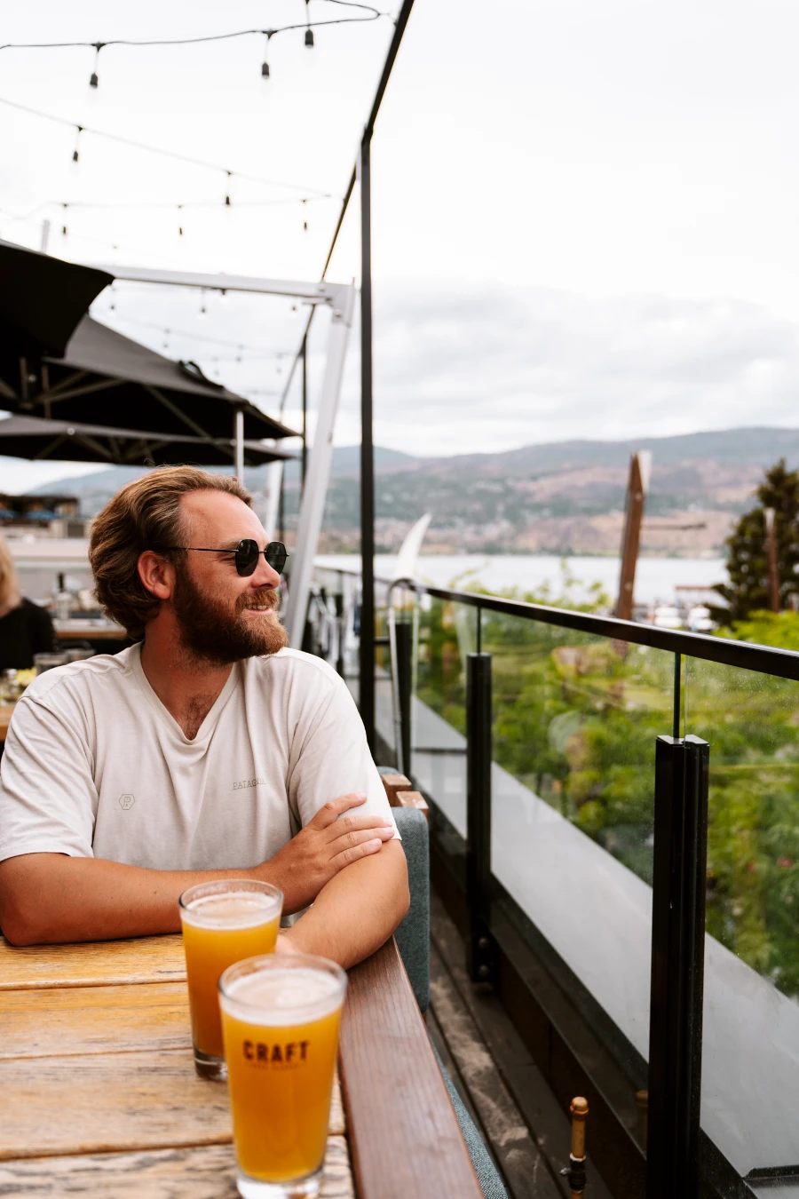 A man sits on a rooftop patio in Kelowna looking out to the side with 2 full pints of hazy beer sitting on the wooden table in front of him