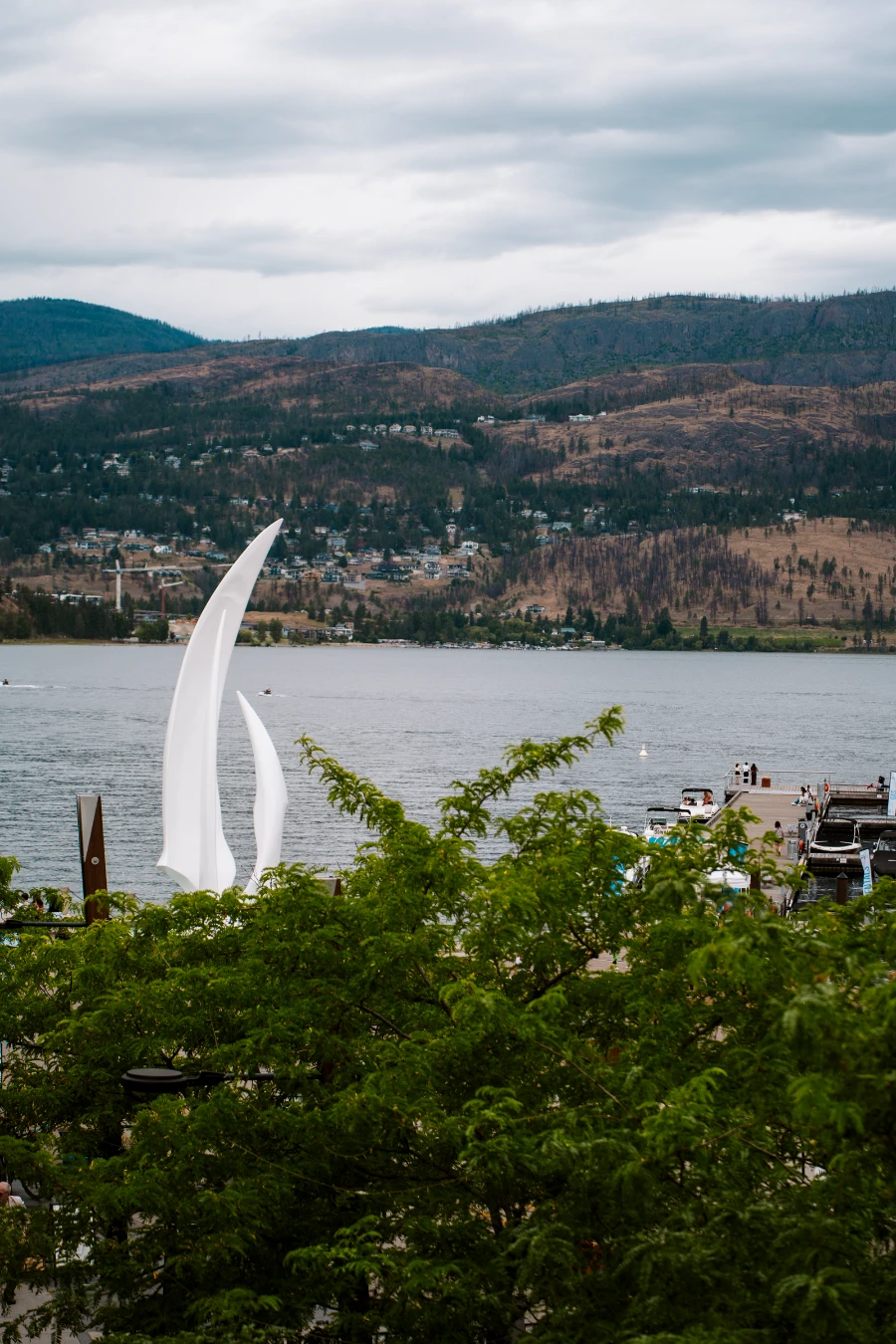 Framed by lush green trees, a white monument towers over the calm waters of Okanagan Lake with rolling hills in the distance