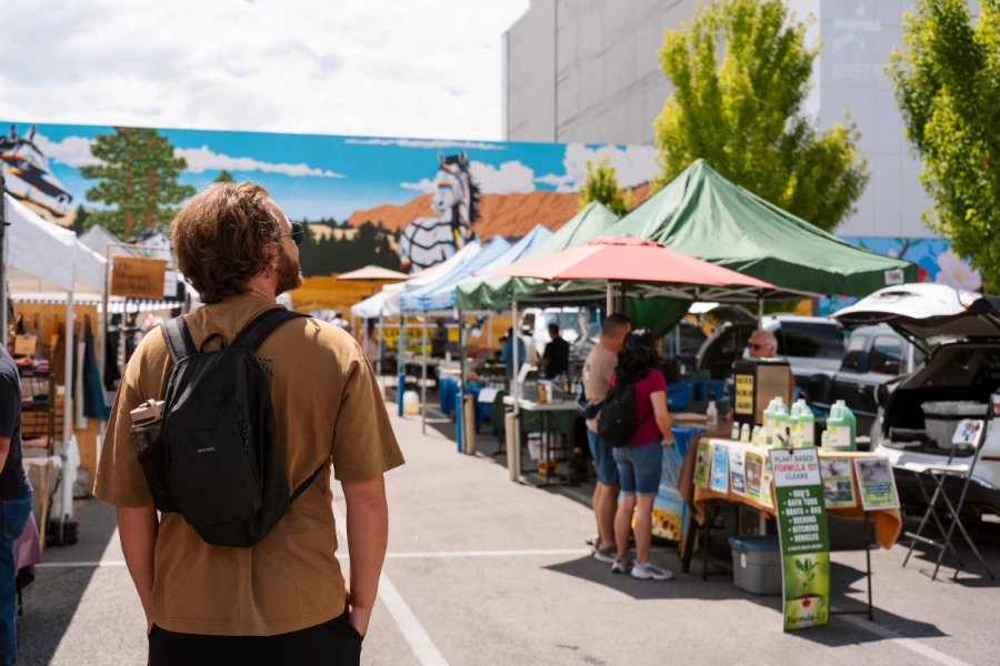 A man with a small black backpack stands looking out to the Kelowna Farmers’ Market and its rows of stalls that line the parking lot, with a large building with an intricate mural painted on it in the distance