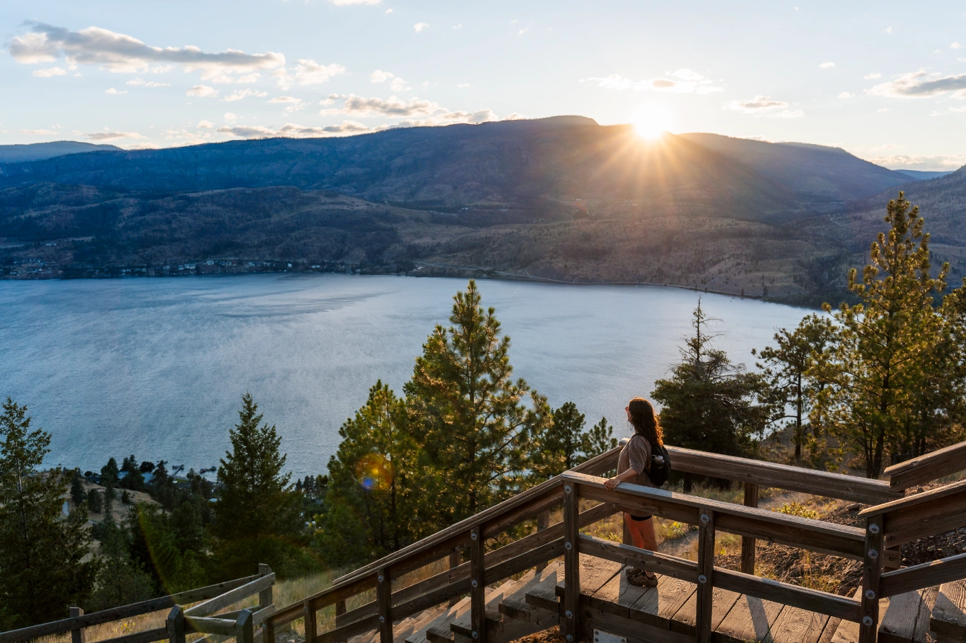 A woman stands on a wooden staircase at the top of Knox Mountain in Kelowna with the sun setting behind a mountain across the lake below her.